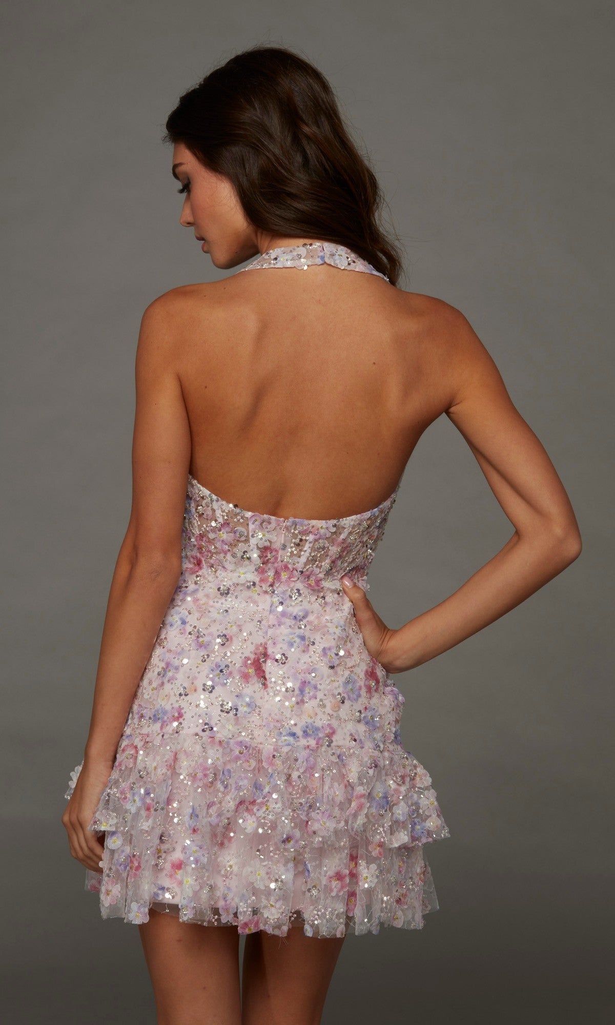 A woman stands with her back to the camera against a plain gray background, modeling the Alyce 30128 short homecoming dress—a pink, backless halterneck style embellished with sequins and finished with a ruffled hem.